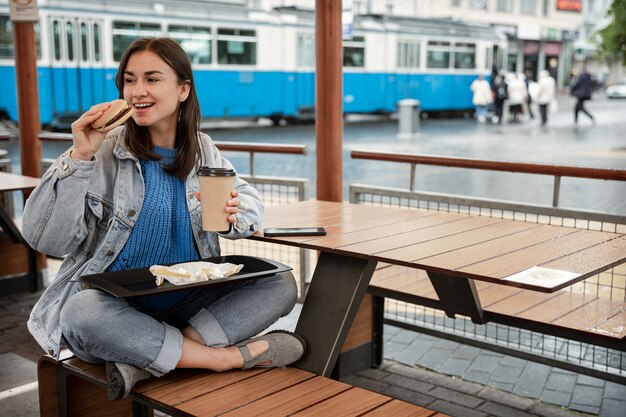 Garota atraente em estilo casual come um hambúrguer com café sentada na esplanada de um café