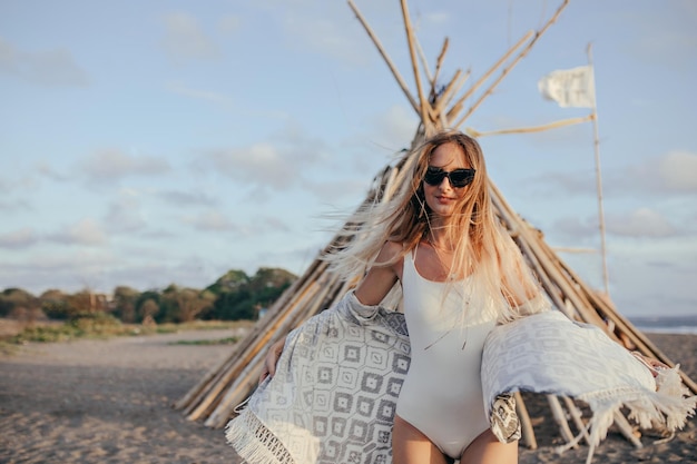 Garota atraente em óculos escuros posando com sorriso interessado na praia retrato ao ar livre de mulher de cabelos compridos feliz usa roupa de banho branca