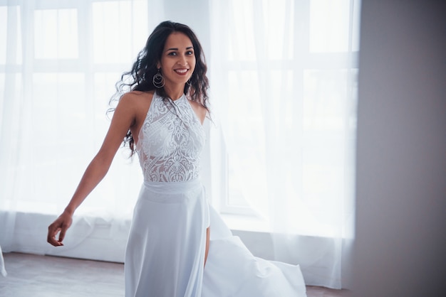 Garota atraente. mulher bonita de vestido branco fica na sala branca com luz do dia através das janelas