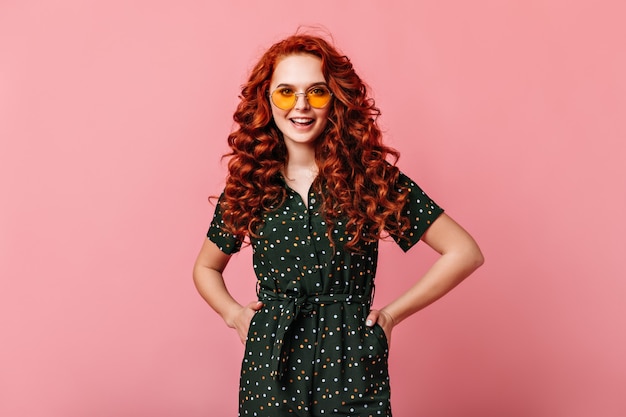 Garota atraente ruiva em pé com as mãos nos quadris. mulher jovem encaracolada feliz em óculos de sol posando em fundo rosa.