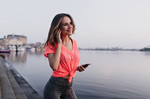Garota bem humorada ouvindo música após o treino. Corredor feminino atraente posando perto do rio com um sorriso.