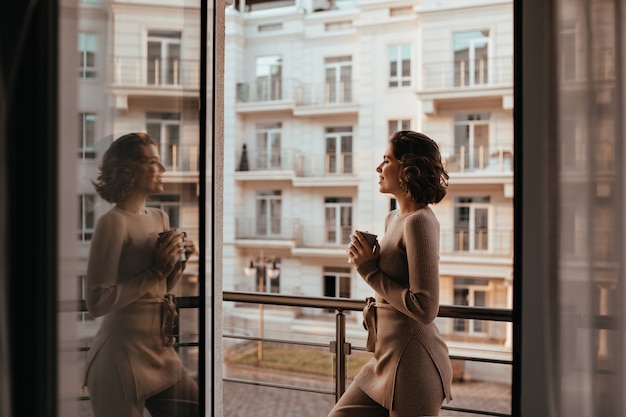 Garota bem torneada e satisfeita bebendo cappuccino e olhando para a cidade. foto de senhora bem-humorada com uma xícara de chá.