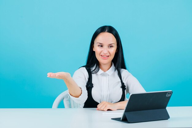 Garota blogueira sorridente está apontando para a esquerda com ahnd sentando-se na frente da câmera do tablet em fundo azul