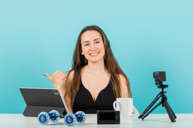 Garota blogueira sorridente está olhando para a câmera apontando para a esquerda com o polegar no fundo azul