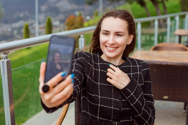 Garota blogueira sorridente está tirando selfie com a câmera traseira do smartphone segurando a mão no peito no fundo da vista da natureza