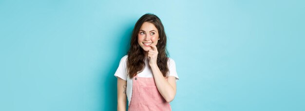 Foto grátis garota boba pensativa com penteado encaracolado fofo mordendo o dedo e olhando para a esquerda no logotipo promocional sorrindo