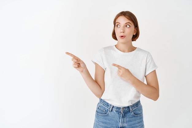 Garota bonita animada com maquiagem natural apontando e olhando para a esquerda divertida diga uau de pé na camiseta contra o fundo branco