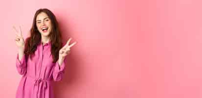 Foto grátis garota bonita positiva em vestido elegante mostrando paz vsign e sorrindo feliz posando perto de fundo rosa