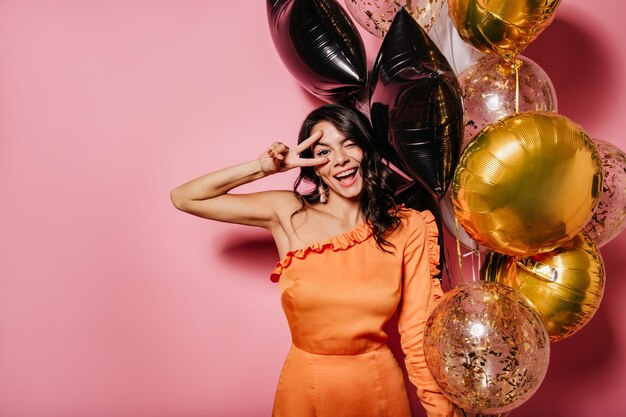 Garota bronzeada despreocupada curtindo festa com sorriso Encantadora mulher latina com balões rindo no fundo rosa