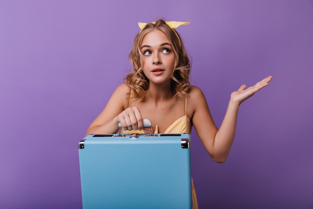 Garota bronzeada posando com mala após a viagem. retrato interior de uma mulher bonita com cabelo loiro em pé violeta.