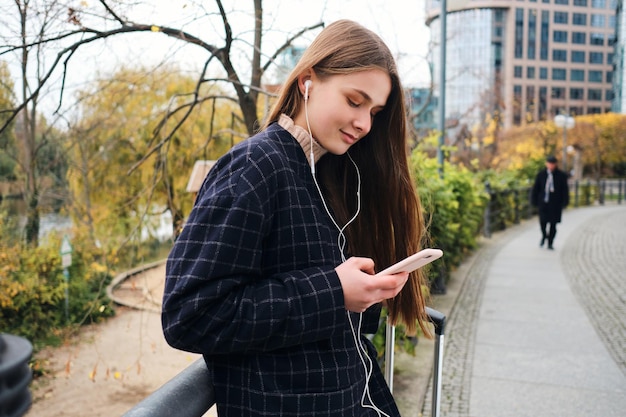 Foto grátis garota casual atraente em fones de ouvido ouvindo música alegremente no celular durante a caminhada no parque da cidade