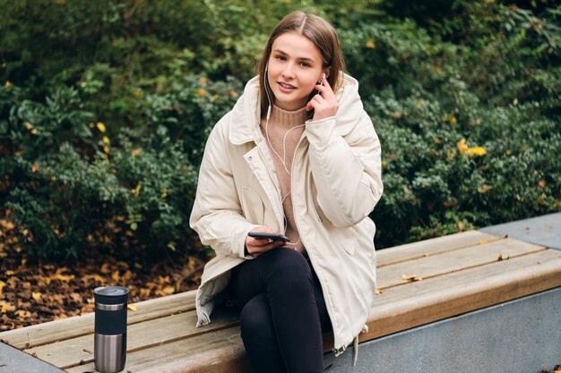 Garota casual atraente olhando alegremente na câmera ouvindo música no celular no parque da cidade