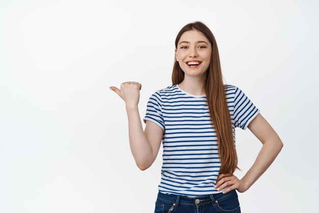 Garota caucasiana feliz rindo, sorrindo e apontando casualmente o dedo para o espaço vazio, banner de venda de compras, de pé sobre fundo branco.