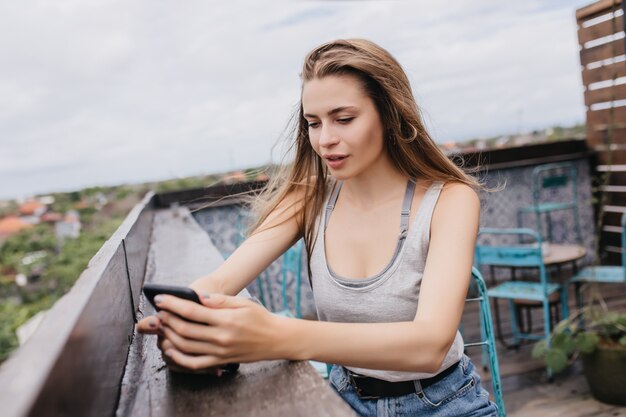 Garota caucasiana inspirada com smartphone passando um tempo no café do telhado. Retrato de uma senhora branca refinada, digitando mensagem em um dia quente de primavera.