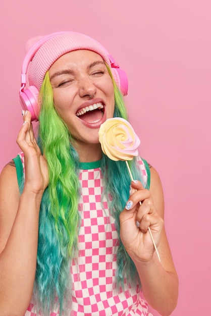 Garota com cabelo comprido tingido segurando deliciosos doces no palito ouve música favorita em fones de ouvido usa vestido casual e chapéu isolado em rosa