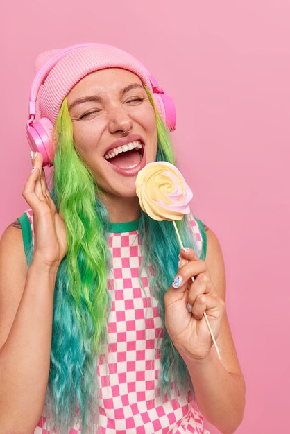 garota com cabelo comprido tingido segurando deliciosos doces no palito ouve música favorita em fones de ouvido usa vestido casual e chapéu isolado em rosa