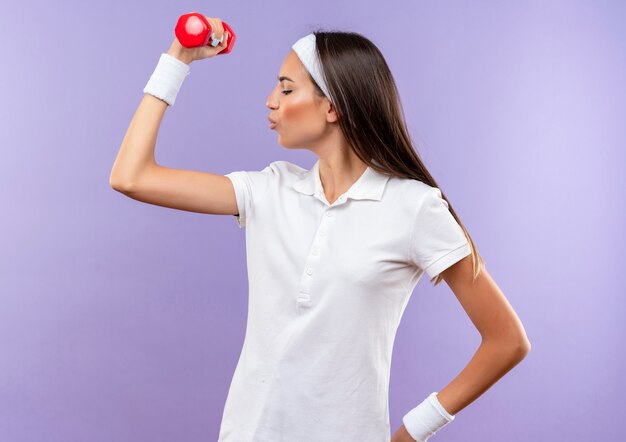Garota confiante e bem esportiva usando bandana e pulseira levantando halteres isolado na parede roxa