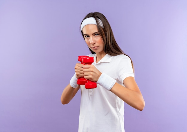 Garota confiante e bem esportiva usando bandana e pulseira segurando halteres isolados na parede roxa com espaço de cópia