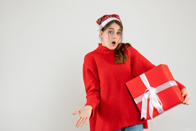 Garota confusa com um chapéu de papai noel segurando um presente de frente