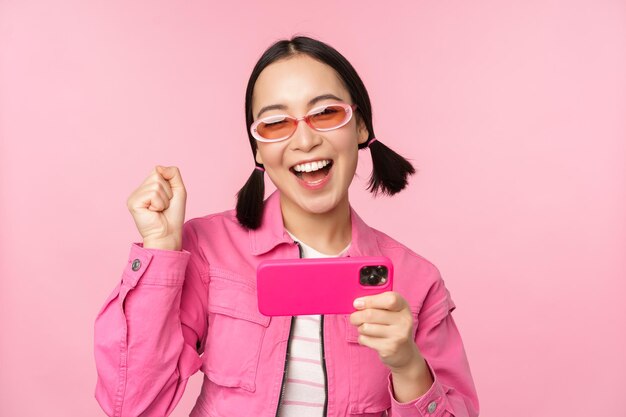 Garota coreana sorridente feliz ganhando no celular olhando para a tela horizontal do smartphone e regozijando-se alcançar o objetivo comemorando em pé sobre fundo rosa