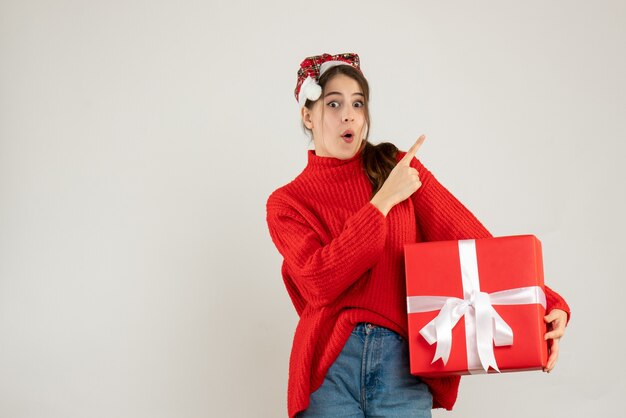 Garota curiosa com chapéu de Papai Noel segurando o dedo no presente e apontando algo em pé