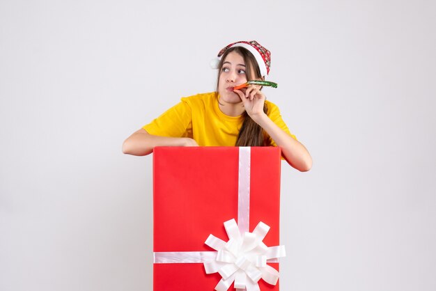 Garota curiosa de frente para o chapéu de papai noel usando o noisemaker atrás de um grande presente de natal