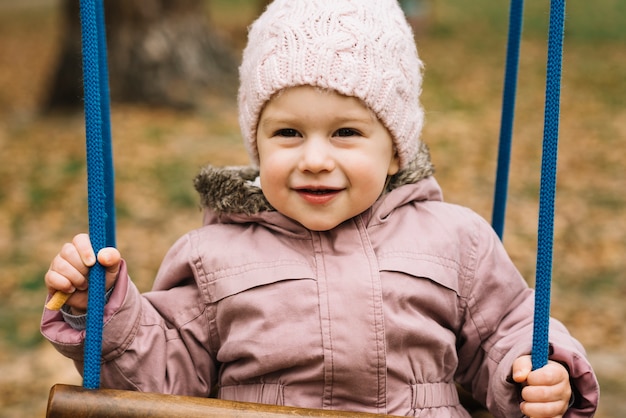 Garota da criança no chapéu de malha balançando no jardim de outono