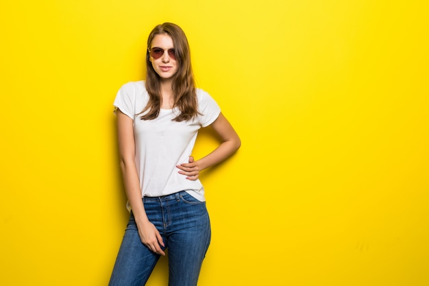 Garota da moda jovem em camiseta branca e jeans azul fica na frente do fundo amarelo do estúdio