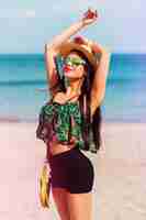 Foto grátis garota de ajuste perfeito na moda óculos de sol legais com corpo bronzeado, posando na praia tropical vestindo blusa de cor brilhante, shorts hight e acessórios de estilo elegante.