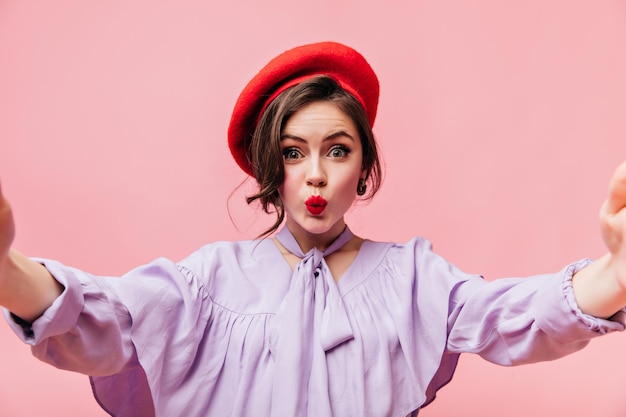 Garota de blusa lilás e boina vermelha manda beijo e tira selfie em fundo rosa