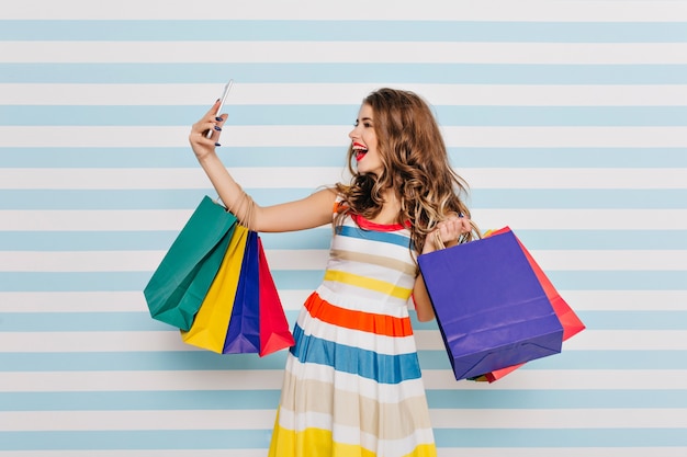 Foto grátis garota de cabelos castanhos inspirada fazendo selfie depois de fazer compras e rir. mulher caucasiana elegante shopaholic segurando sacolas e tirando uma foto de si mesma.