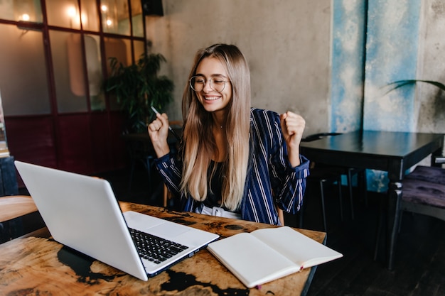 Garota de cabelos compridos animada, se divertindo durante o trabalho com o computador. Foto interna de freelancer feminino sorridente usando laptop em um café aconchegante.