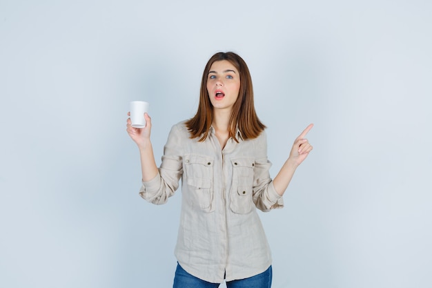 garota de camisa segurando um copo plástico de café, apontando para o canto superior direito e parecendo inteligente.