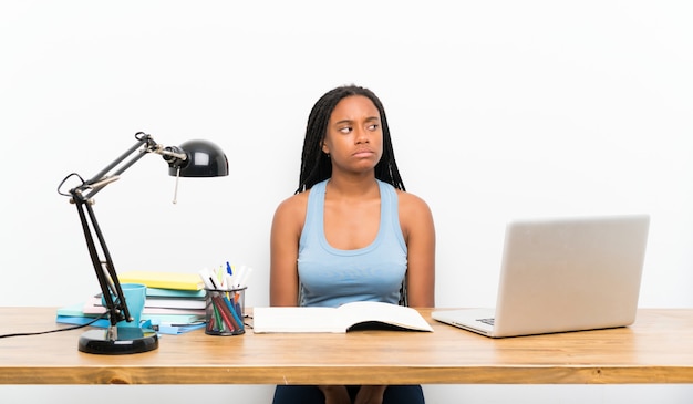 Garota de estudante adolescente americano africano com cabelo longo trançado no seu local de trabalho, fazendo dúvidas gesto olhando lado