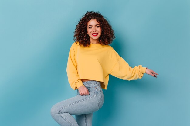 Garota de olhos azuis com lábios brilhantes e sorriso branco como a neve está posando no espaço azul. Senhora de suéter amarelo e jeans skinny dançando.