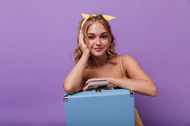 Garota de olhos escuros satisfeita posando com uma valise azul. Mulher bonita entusiasmada sentada na violeta com um sorriso gentil.