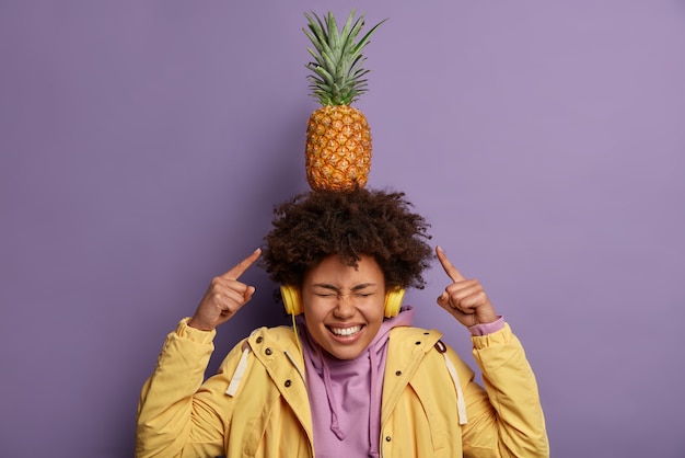 Garota de pele escura radiante carrega abacaxi exótico na cabeça e ri positivamente ouve música em fones de ouvido estéreo vestida casualmente indo comer frutas frescas, brinca, isolado sobre parede violeta