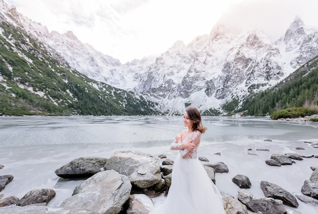 Garota de sorriso frio vestida com o vestido de noiva está de pé perto do lago das montanhas congeladas no inverno