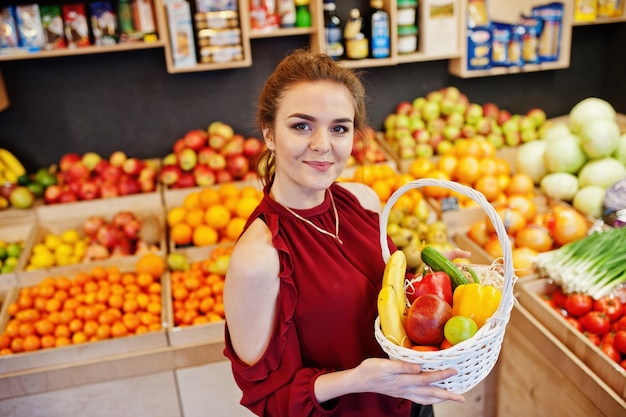 Garota de vermelho segurando diferentes frutas e legumes na cesta na loja de frutas
