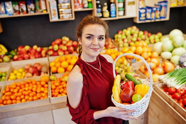 Garota de vermelho segurando diferentes frutas e legumes na cesta na loja de frutas