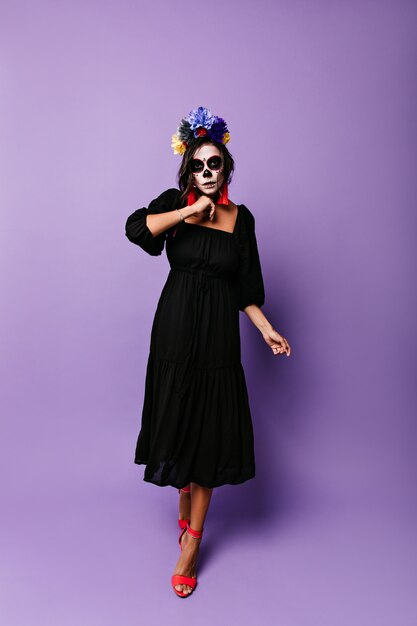 Garota de vestido preto midi caminha contra a parede roxa. Modelo com máscara de caveira em poses de rosto para foto de Halloween.