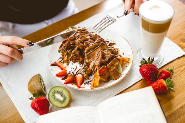 Garota de vista lateral comendo waffles de chocolate de sobremesa com frutas com café com leite