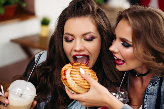 Garota deslumbrante com maquiagem alimentando sua amiga com hambúrguer no café.