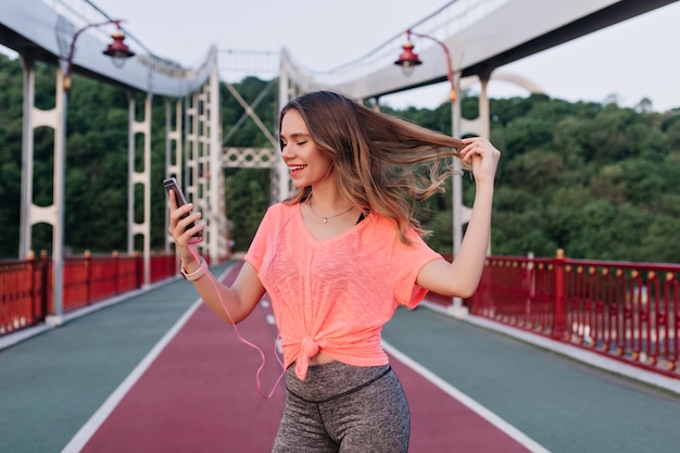 Garota despreocupada brincando com seu cabelo enquanto tirava uma foto de si mesma no estádio. Mulher caucasiana incrível fazendo selfie com smartphone.