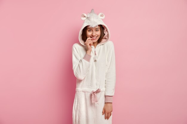 Garota despreocupada com aparência europeia, usa traje branco macio de kigurumi, mantém o dedo da frente nos lábios, encosta na parede rosa, tem tempo livre em casa. Pessoas, emoções, conceito de estilo de vida