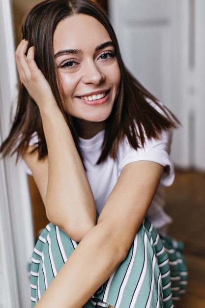 Garota despreocupada com grandes olhos castanhos rindo enquanto está sentado em casa. Foto interna de magnífica senhora morena de pijama relaxante no fim de semana.