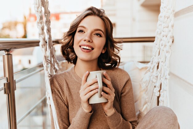 Garota despreocupada com maquiagem marrom, bebendo chá na varanda. Foto de uma agradável mulher morena com vestido de malha, desfrutando do café.