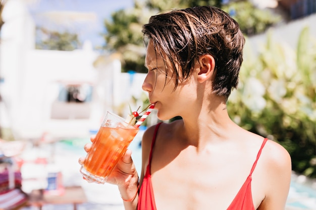 Garota despreocupada com o corte de cabelo da moda, bebendo coquetel de frutas no resort.