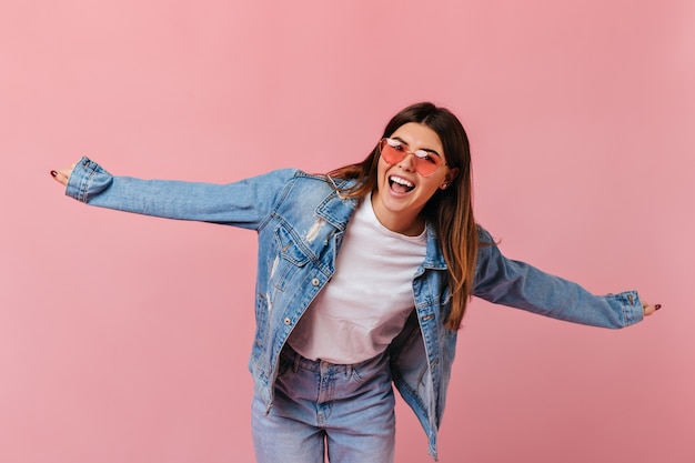 Garota despreocupada em óculos de sol rindo sobre fundo rosa. foto de estúdio de jovem animada em jeans se divertindo.