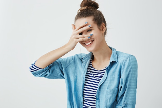 Garota dizendo palavras tolas e fica envergonhada. foto interior de bonita mulher esbelta em vestuário elegante, cobrindo os olhos com a mão, mas espreitando, sorrindo amplamente, expressando emoções positivas